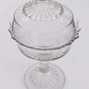 May include: Clear glass butter dish with a lid. The dish has a pedestal base and a scalloped edge design. The lid has a decorative knob and a matching scalloped pattern. The dish is approximately 6 inches tall.