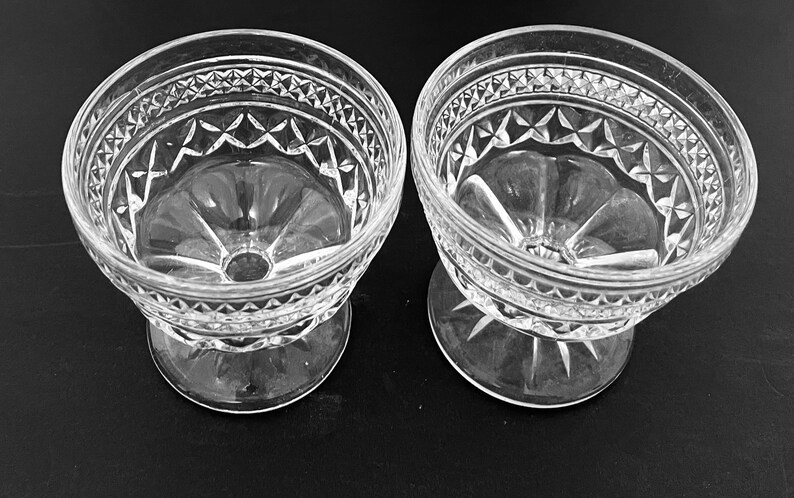May include: Two clear glass dessert bowls with a decorative diamond and starburst pattern. The bowls have a pedestal base and a wide rim with a repeating geometric design. The bowls are likely vintage and suitable for serving ice cream or other desserts.