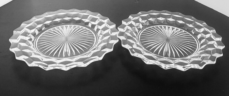 K&ouml;nnte beinhalten: Zwei klare Glasplatten mit einem Sternmuster in der Mitte und einem rautenf&ouml;rmigen Rand. Die Teller haben einen gewellten Rand und werden auf einer dunklen Oberfl&auml;che pr&auml;sentiert.