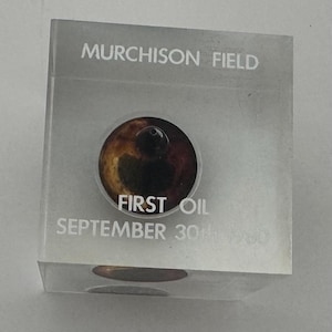 May include: A clear, rectangular block with the text "Murchison Field" and "First Oil September 30th 1980" printed on it. A circular emblem with brown and black tones is centered on the block.