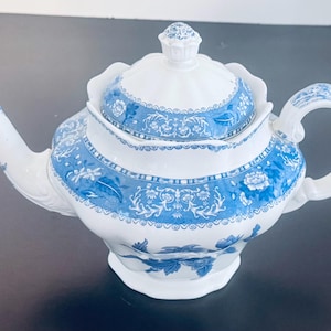 Puede incluir: Tetera de cerámica blanca con un diseño floral azul. La tetera tiene un pico curvo, un asa redondeada y una tapa con un adorno decorativo. El patrón azul incluye diseños florales y de volutas, creando un aspecto clásico y elegante.