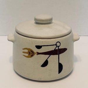 Puede incluir: Un tarro de cerámica color crema con tapa y dos asas pequeñas. El tarro presenta un diseño marrón y amarillo de un tenedor y cucharas. La tapa tiene un pequeño pomo. El tarro es probablemente para uso en la cocina.