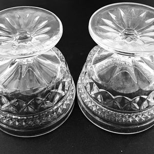 May include: Two clear, cut-glass dessert bowls with a decorative pattern. The bowls are inverted, showing the detailed design of the base and the starburst pattern on the foot. The bowls are likely made of crystal.