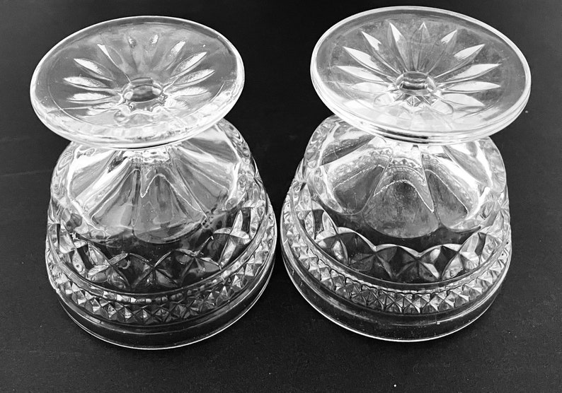 May include: Two clear, cut-glass dessert bowls with a decorative pattern. The bowls are inverted, showing the detailed design of the base and the starburst pattern on the foot. The bowls are likely made of crystal.