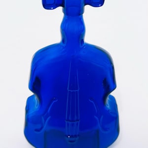 May include: A cobalt blue glass bottle shaped like a violin. The bottle has a long neck with two rounded handles and a detailed design on the front, resembling the instrument's features. The bottle is made of transparent glass.