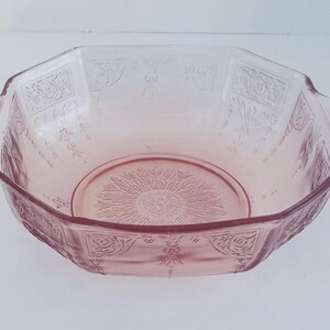 May include: A pink, octagonal glass bowl with handles. The bowl features a floral and geometric pattern around the rim and a sunburst design on the base. The bowl is made of a translucent pink glass.