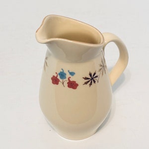 May include: Cream-colored ceramic pitcher with a curved spout and handle. The pitcher is decorated with red, blue, and gold floral and starburst designs. A vintage-style creamer or small water pitcher.