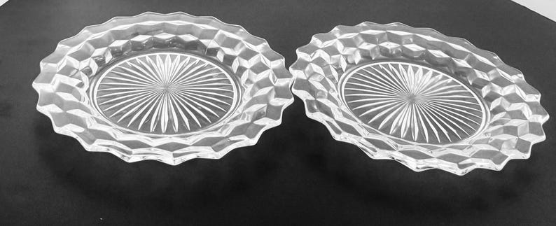 K&ouml;nnte beinhalten: Zwei klare Glasplatten mit einem Sternmuster in der Mitte und einem rautenf&ouml;rmigen Rand. Die Teller haben einen gewellten Rand und liegen auf einer dunklen Oberfl&auml;che. Diese Vintage-Teller eignen sich zum Servieren von Vorspeisen oder Desserts.