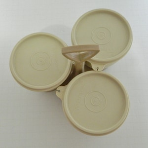 Puede incluir: Un juego de recipientes Tupperware beige con tres compartimentos redondos y tapas a juego. Las tapas tienen un diseño circular en el centro y la palabra "Tupperware" está impresa en cada tapa. Un asa beige conecta los compartimentos.