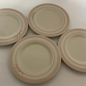 May include: Four off-white ceramic plates with a light brown double border and a decorative flourish. The plates are arranged on a white surface. The plates are likely for serving food.