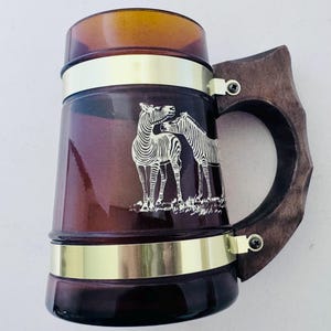 Puede incluir: Un jarro de cerveza vintage de vidrio marrón con un asa de madera y bandas metálicas doradas. El jarro presenta una ilustración blanca de dos cebras. El asa está sujeta con pequeños soportes metálicos.