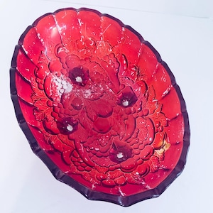 May include: A red oval-shaped glass serving bowl with a dark rim. The bowl features a floral design with embossed leaves and flowers. The bowl is on a white background.