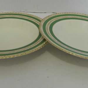 May include: Two oval serving platters. The plates have a cream-coloured centre, a green band, and a gold rope-like border. The plates are displayed on a white surface.