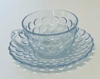 Puede incluir: Un juego de taza y platillo de té de vidrio azul claro. La taza y el platillo tienen un patrón texturizado similar a burbujas. La taza tiene un asa y se asienta sobre un platillo a juego, creando una estética delicada y elegante. Perfecto para servir té o café.