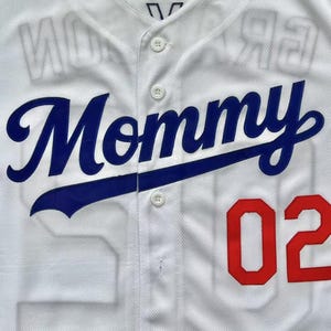 Pode incluir: Uma camisa de beisebol branca com bot&otilde;es, apresentando a palavra Mommy em letras cursivas azuis no peito. O n&uacute;mero 02 est&aacute; impresso em vermelho no lado inferior direito. O tecido tem um padr&atilde;o de malha texturizada e uma etiqueta L na gola.