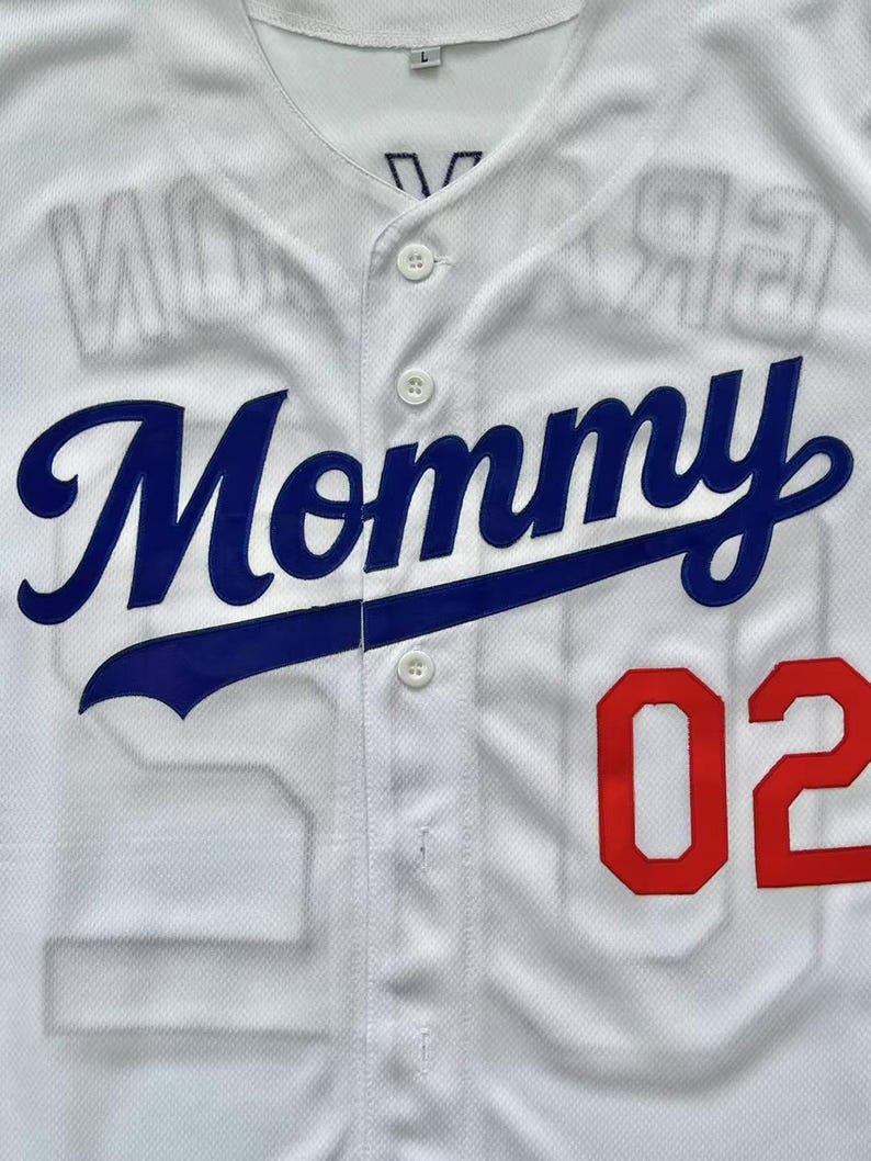 Pode incluir: Uma camisa de beisebol branca com bot&otilde;es, apresentando a palavra Mommy em letras cursivas azuis no peito. O n&uacute;mero 02 est&aacute; impresso em vermelho no lado inferior direito. O tecido tem um padr&atilde;o de malha texturizada e uma etiqueta L na gola.