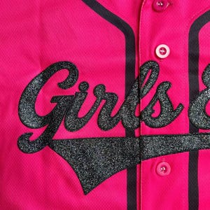 May include: A bright pink baseball jersey with the word "Girls" in large, glittery black letters across the front. The jersey has black stripes along the button placket and collar, with matching black trim. Pink buttons secure the front.