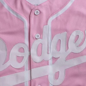 May include: Pink baseball jersey with white accents and the word "Dodgers" in white lettering. The jersey has a button-up front and a V-neck collar. The fabric has a textured appearance.
