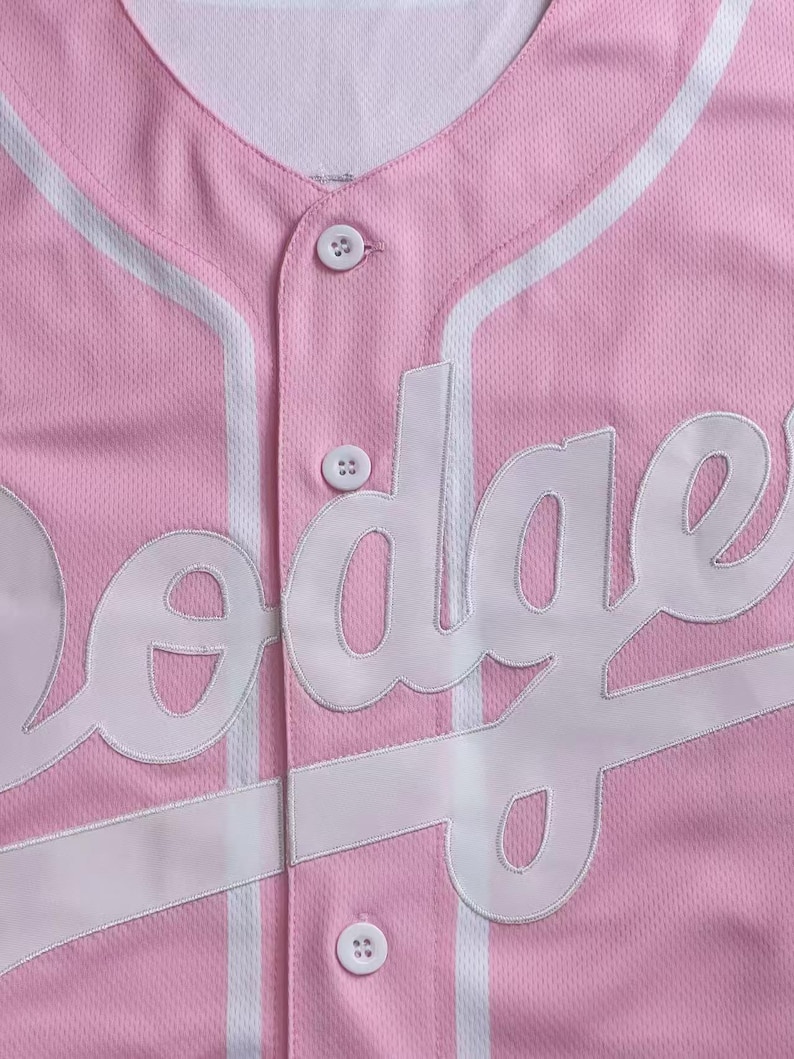 May include: Pink baseball jersey with white accents and the word "Dodgers" in white lettering. The jersey has a button-up front and a V-neck collar. The fabric has a textured appearance.