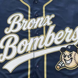 Pode incluir: Uma camisa de beisebol azul-marinho com bot&otilde;es, apresentando o texto branco "Bronx Bombers" em fonte cursiva com contorno dourado. Um gr&aacute;fico de um personagem de beisebol em desenho animado usando um bon&eacute; com logo est&aacute; na parte inferior direita.