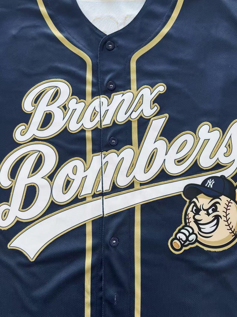 Pode incluir: Uma camisa de beisebol azul-marinho com bot&otilde;es, apresentando o texto branco "Bronx Bombers" em fonte cursiva com contorno dourado. Um gr&aacute;fico de um personagem de beisebol em desenho animado usando um bon&eacute; com logo est&aacute; na parte inferior direita.