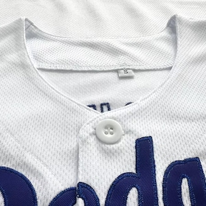 Pode incluir: Uma camisola de basebol branca em tecido de malha texturizado, com um &uacute;nico bot&atilde;o branco na gola e letras bordadas em azul escuro na parte da frente. Uma pequena etiqueta branca com a letra S &eacute; vis&iacute;vel no interior da gola.