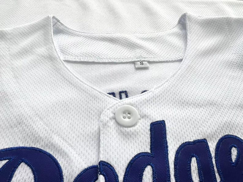 Pode incluir: Uma camisola de basebol branca em tecido de malha texturizado, com um &uacute;nico bot&atilde;o branco na gola e letras bordadas em azul escuro na parte da frente. Uma pequena etiqueta branca com a letra S &eacute; vis&iacute;vel no interior da gola.