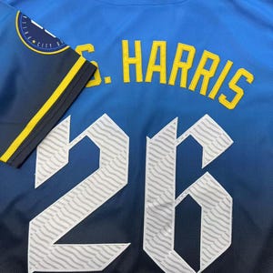 Pode incluir: Uma camisola desportiva azul com o nome S. HARRIS em letras amarelas acima do n&uacute;mero 26 em branco com um padr&atilde;o ondulado. A manga &eacute; azul-escura com uma risca amarela e um emblema circular com o texto CITY EDITION.