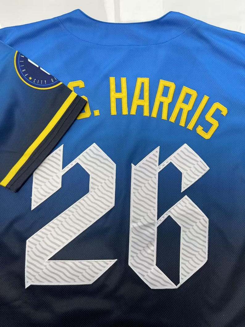 Pode incluir: Uma camisola desportiva azul com o nome S. HARRIS em letras amarelas acima do n&uacute;mero 26 em branco com um padr&atilde;o ondulado. A manga &eacute; azul-escura com uma risca amarela e um emblema circular com o texto CITY EDITION.
