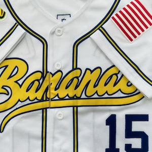 Pode incluir: Uma camisa de beisebol branca com a palavra Bananas em letra amarela com contorno azul-escuro no peito. A camisa tem debrum azul-escuro, bot&otilde;es brancos, o n&uacute;mero 15 em azul-escuro na frente e um emblema com listras vermelhas e brancas.