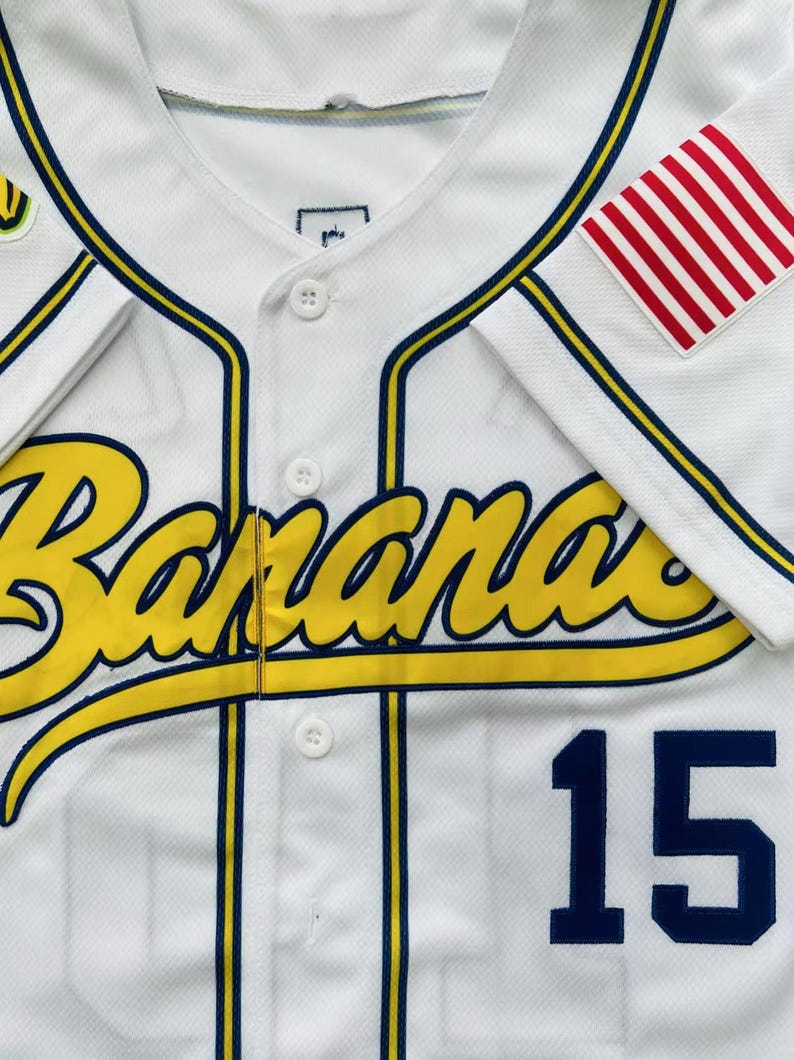 Pode incluir: Uma camisa de beisebol branca com a palavra Bananas em letra amarela com contorno azul-escuro no peito. A camisa tem debrum azul-escuro, bot&otilde;es brancos, o n&uacute;mero 15 em azul-escuro na frente e um emblema com listras vermelhas e brancas.