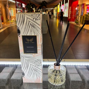 May include: A diffuser bottle with black reeds and a white and grey box with a palm leaf design. The box has the text "Parlour + More" and "Vanilla Caramel" on it. The bottle is clear glass with a gold lid and a textured design. The bottle is sitting on a white surface.