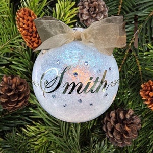 May include: A white, glittery Christmas ornament with a gold ribbon and the name "Smith" written in cursive on the front.