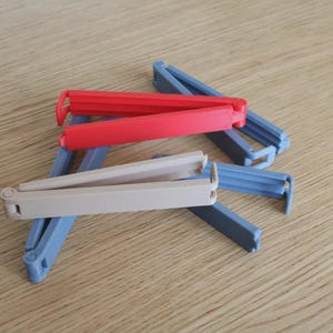 May include: A collection of colorful plastic bag clips in red, beige, and blue. The clips are designed to seal bags and keep contents fresh. They are arranged on a light brown wooden surface.