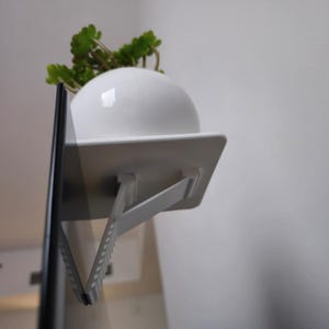 May include: White plastic shelf with a plant in a white pot attached to a black television screen. The shelf has a white bracket that folds down to support the shelf.