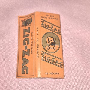 May include: A vintage pack of Zig-Zag rolling papers. The pack is orange with green and black text, including "SLOW BURNING" and "MADE IN FRANCE." The pack features a circular illustration of a man smoking. The pack contains 75 leaves.