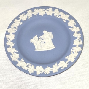 May include: A round, light blue plate with a white relief design. The plate features a white border of leaves and vines. In the center is a white relief of figures. The plate is likely ceramic or porcelain.