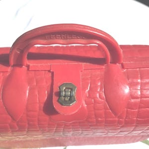 May include: A red, vintage-style cosmetic case with a textured, crocodile-like pattern. The case features a handle with the word "PEERLESS" embossed on it, a metal clasp, and a rounded shape.