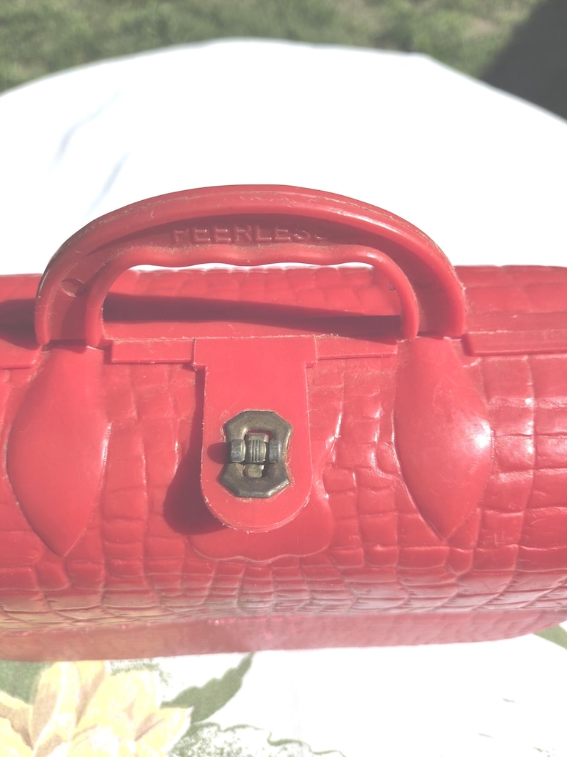 May include: A red, vintage-style cosmetic case with a textured, crocodile-like pattern. The case features a handle with the word "PEERLESS" embossed on it, a metal clasp, and a rounded shape.
