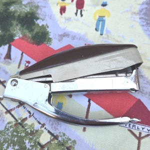 May include: A vintage stapler with a brown top and silver metal base. The stapler is resting on a colorful fabric with a train and town scene. The stapler's design is classic and functional.