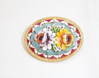 Puede incluir: Un broche ovalado con un diseño de mosaico y un borde de metal trenzado en tono dorado. El centro muestra patrones florales en tonos amarillo, naranja, blanco y azul claro, rodeados por un borde decorativo de azulejos geométricos rojos y blancos.