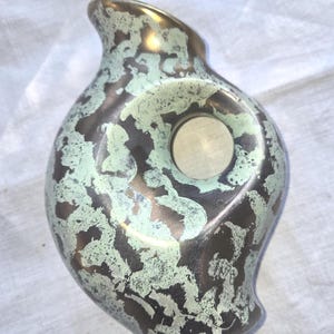 May include: A decorative vase with a unique shape and a mottled finish. The vase features a bronze base with a green and brown pattern, and a gold-colored rim. A circular indentation is visible on the side of the vase.
