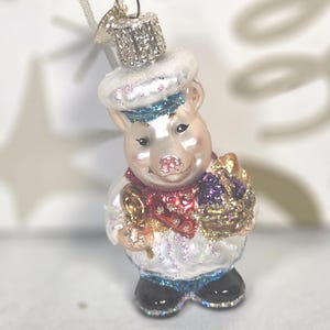May include: A glass pig ornament wearing a white hat with blue glitter, a red scarf, and black shoes. The pig holds a gold-toned basket filled with purple and gold glittered items. A silver metal cap with a hanging loop sits atop the ornament.