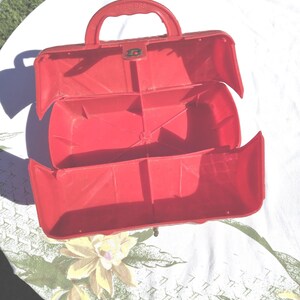May include: Open red plastic toolbox with a handle and a clasp. The toolbox is open, revealing interior compartments. The word "HANDLESS" is embossed on the handle. The toolbox is sitting on a floral patterned tablecloth.