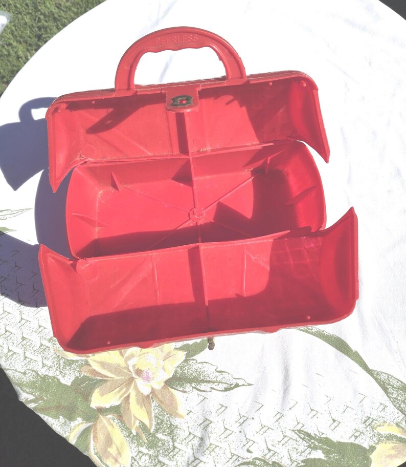 May include: Open red plastic toolbox with a handle and a clasp. The toolbox is open, revealing interior compartments. The word "HANDLESS" is embossed on the handle. The toolbox is sitting on a floral patterned tablecloth.