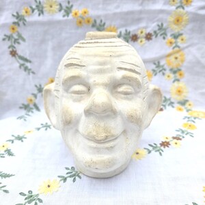 May include: A ceramic sculpture of a human head with a weathered off-white finish. The face features closed eyes, a prominent nose, and a gentle smile. The top of the head has a small circular opening, and the ears are large and rounded.