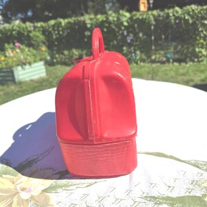 May include: A vintage red plastic lunchbox with a handle. The lunchbox has a rectangular shape with a textured bottom and a smooth, rounded top. The handle is arched and attached to the top of the lunchbox.