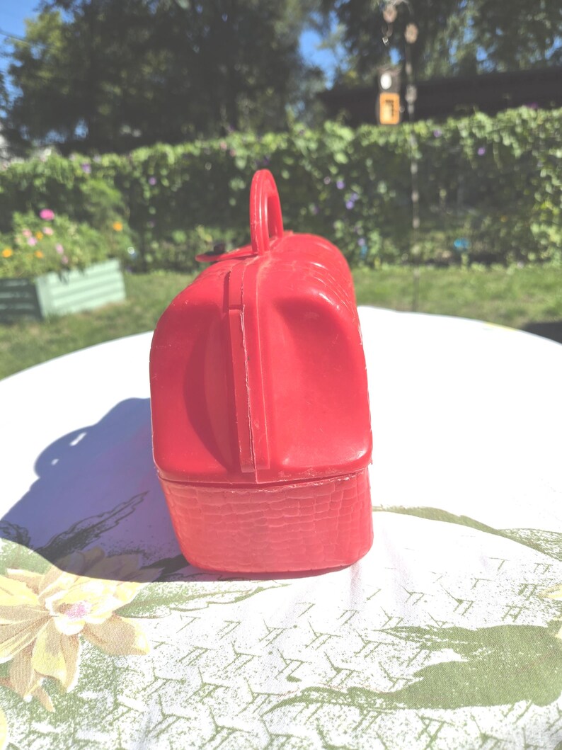 May include: A vintage red plastic lunchbox with a handle. The lunchbox has a rectangular shape with a textured bottom and a smooth, rounded top. The handle is arched and attached to the top of the lunchbox.