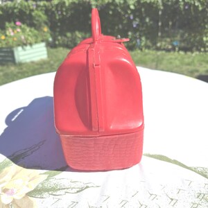 May include: A vintage red lunchbox with a handle and a basket weave design on the bottom. The lunchbox is made of plastic and has a clasp closure. The handle is arched and attached to the top of the lunchbox.