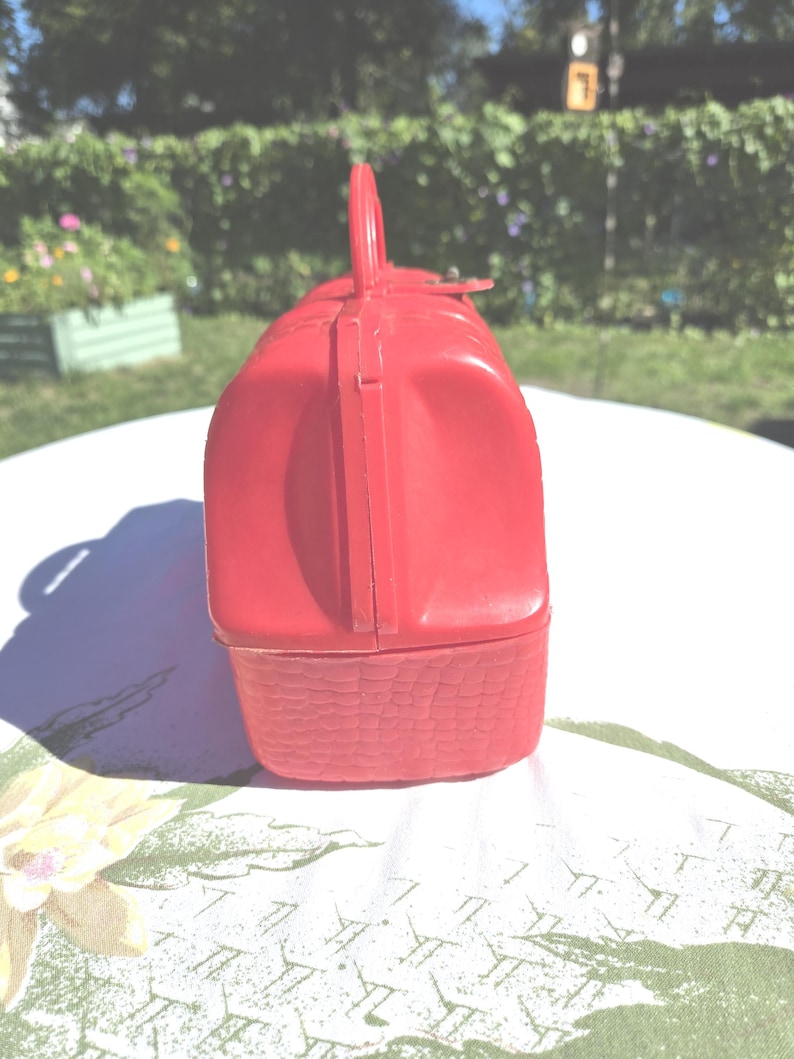 May include: A vintage red lunchbox with a handle and a basket weave design on the bottom. The lunchbox is made of plastic and has a clasp closure. The handle is arched and attached to the top of the lunchbox.